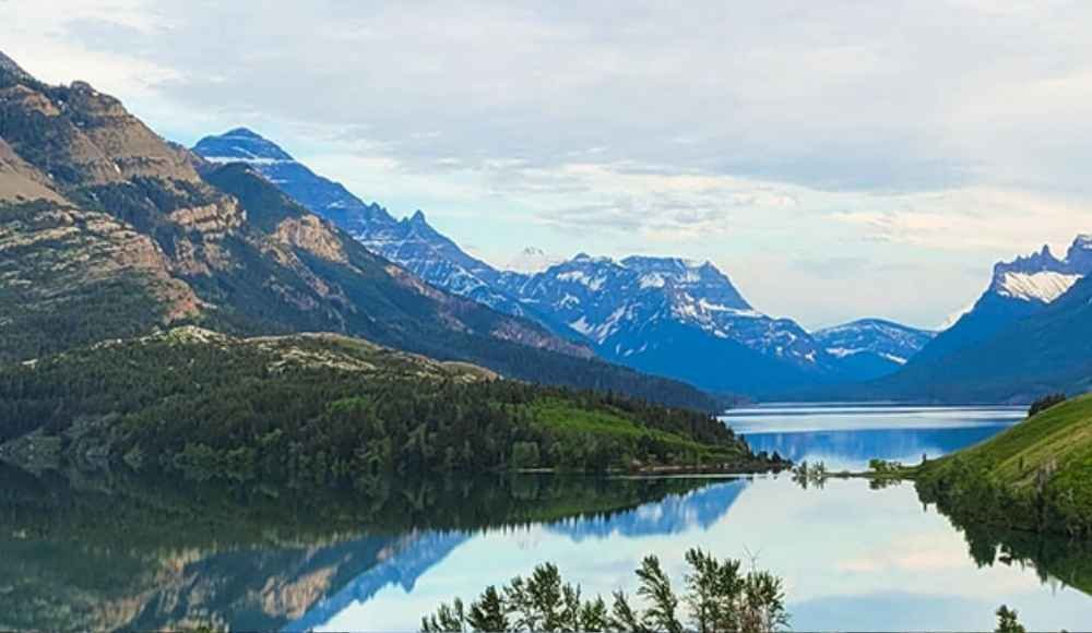 A calm lake reflects surrounding forested hills and snow-capped mountains under a cloudy sky, capturing one of the breathtaking wonders in Alberta.