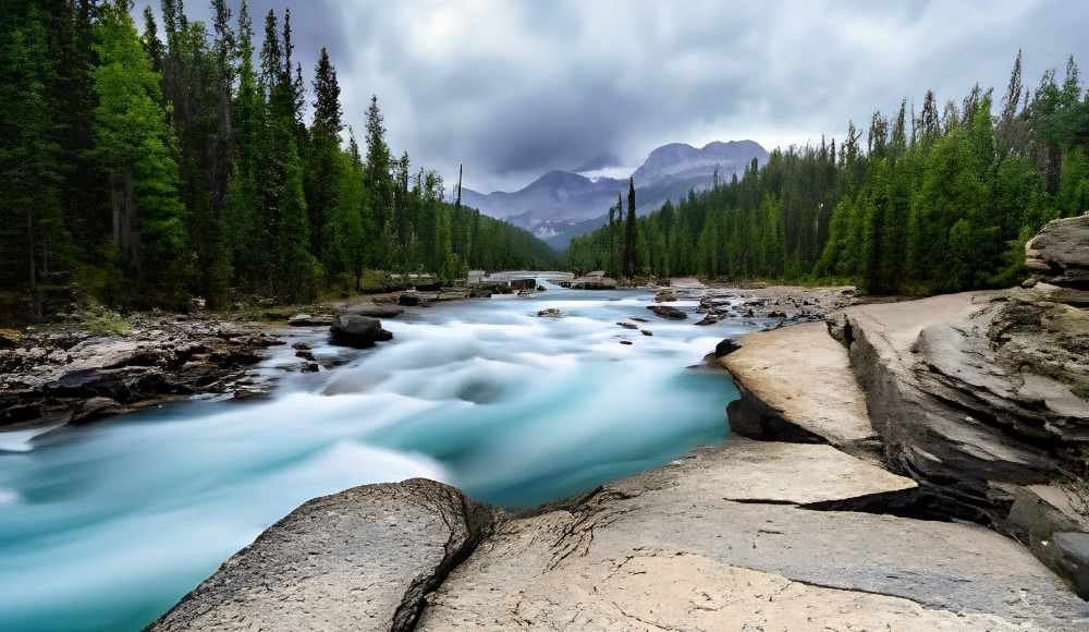 A fast-flowing river winds through a rocky landscape surrounded by dense evergreen forest, set against mountains and a cloudy sky—one of the hidden gems in Alberta for those seeking untamed beauty.