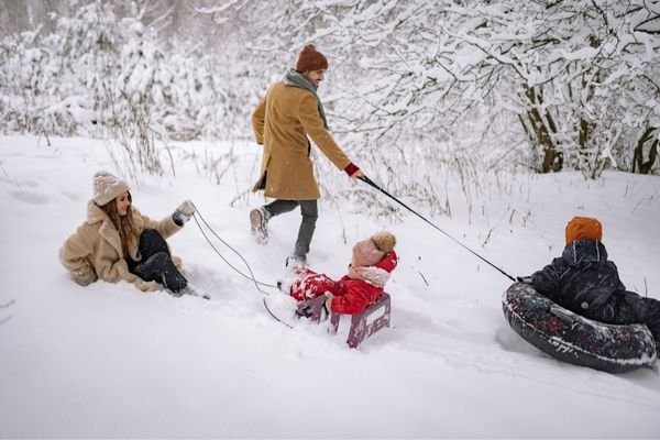 Two adults and two children play in the snow; one adult pulls a child on a sled and another on an inflatable tube, while the second adult sits on the ground.