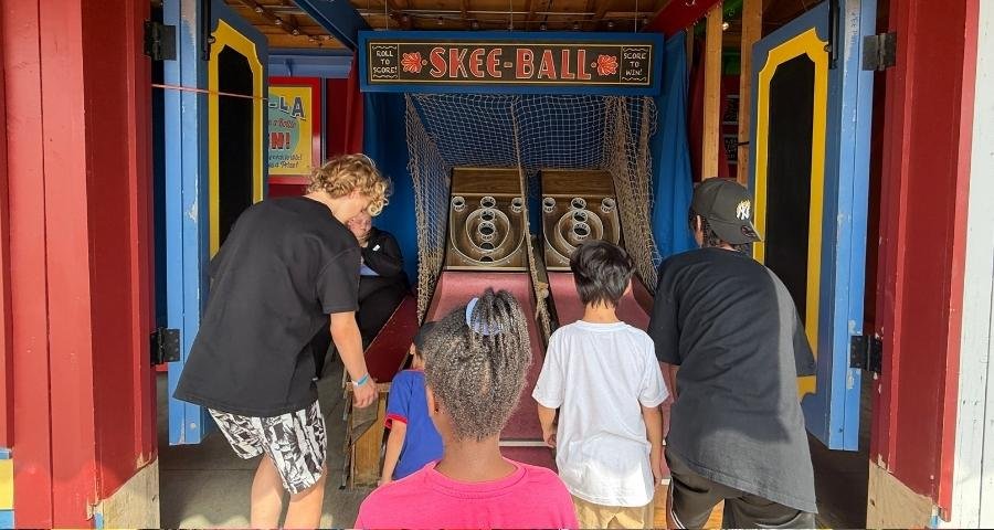 Four children stand in front of a skee-ball arcade game at Fort Edmonton Park, with two playing and two watching. The scene is set inside a colorful booth.