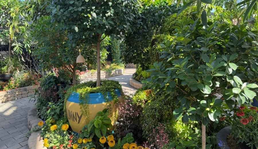 A paved garden path curves past a large yellow-and-blue pot labeled “HUNNY,” surrounded by trees, flowers, and dense greenery reminiscent of the lush displays found at the Muttart Conservatory.