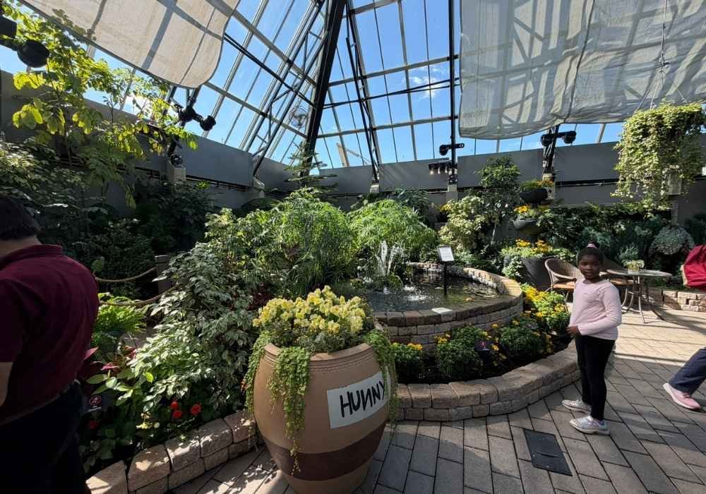 A greenhouse garden with various plants, a central fountain, a large vase labeled “HUMAN,” and people observing the surroundings under a glass ceiling, reminiscent of the lush displays found at the Muttart Conservatory.