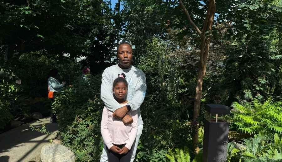 A man stands behind a young girl, resting his arms on her shoulders. They are outdoors at the Muttart Conservatory, surrounded by lush green plants and trees in bright daylight.