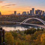 People stand on an overlook facing a river, arched bridge, and city skyline at sunset, with autumn trees in the foreground.